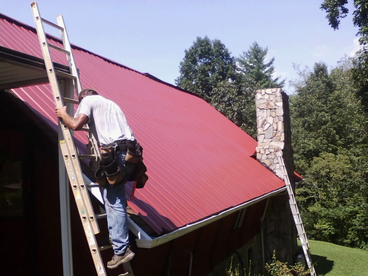 Roofer on a ladder beside a red metal roof and stone chimney — Soffit & Fascia Repair by Roofing Brothers in Cambria